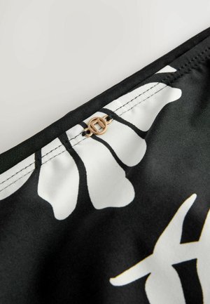 Schwarzer Stoff mit weißen floralen Mustern und einem goldenen, runden Hardware-Detail oben. Glatte Textur mit sichtbaren Kontraststichen.