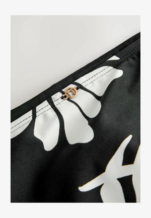 Schwarzer Stoff mit weißen floralen Mustern und einem goldenen, runden Hardware-Detail oben. Glatte Textur mit sichtbaren Kontraststichen.