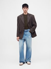 Άνδρας που φορά καφέ oversized σακάκι, σκούπε πράσινο πουλόβερ, φαρδιά ανοιχτό μπλε τζιν και λευκά παπούτσια, όρθιος μπροστά σε απλό ανοιχτό φόντο.