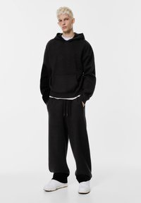 Felpa nera con cappuccio e tasca frontale, sopra una t-shirt bianca, abbinata a pantaloni sportivi neri e sneakers bianche, con un taglio rilassato e una texture morbida.