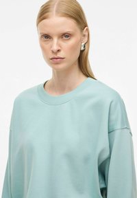 Donna con capelli biondi lisci, che indossa grandi orecchini a cerchio in argento e una felpa oversize azzurra, su uno sfondo bianco semplice.