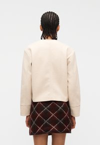 Άτομο με πλεξουδάκια, που φοράει μια μπεζ μακρυμάνικη ζακέτα και μια φούστα με διαμάντια σε κόκκινο, μαύρο και άσπρο χρώμα, γυρισμένο με την πλάτη.