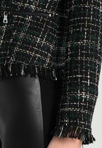 Giacca in tweed con zip, a fantasia scozzese nei colori nero, verde, bianco e giallo, caratterizzata da orlo e polsini frangiati, indossata con pantaloni in pelle nera.