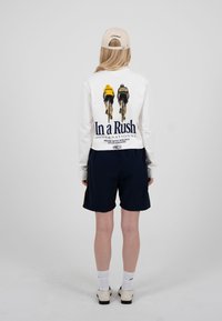 Personne portant une chemise blanche à manches longues avec des cyclistes et le texte "In a Rush" au dos, un short noir, des chaussettes blanches, une casquette beige et des chaussures blanches.