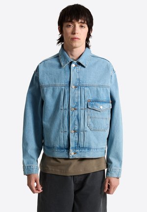 En ung person med mørkt hår iført en lyseblå denimjakke over en brun skjorte og mørke bukser, stående foran en ensfarvet baggrund.