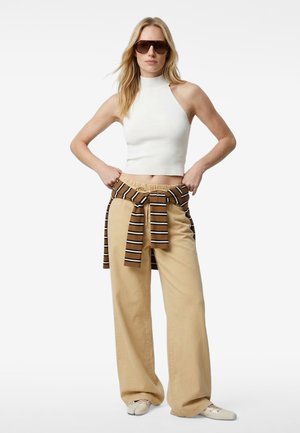 Femme portant un crop top blanc sans manches, un pantalon beige à jambes larges, un pull marron rayé noué à la taille, des chaussures beiges et des lunettes de soleil foncées.
