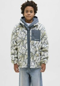 Giacca in pile con motivo floreale in bianco, verde e grigio; chiusura con zip, tasca sul petto grigia e design con cappuccio. Indossata sopra una camicia bianca.