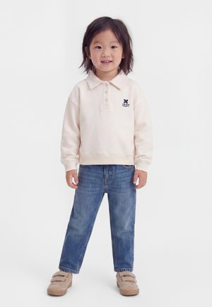 Kind staat en lacht, draagt een crèmekleurig poloshirt met lange mouwen, blauwe spijkerbroek en beige sneakers met klittenbandsluiting tegen een witte achtergrond.