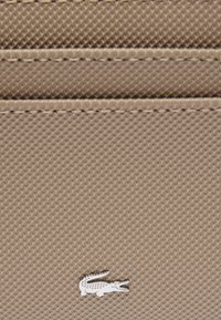 Beige textuur portefeuille met horizontale kaartsleuven en een zilveren krokodillenlogo. Heeft gestikte randen en een gladde afwerking.