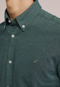 Camisa de botones verde texturizada con cuello, que presenta botones blancos y un pequeño logotipo bordado en el pecho.