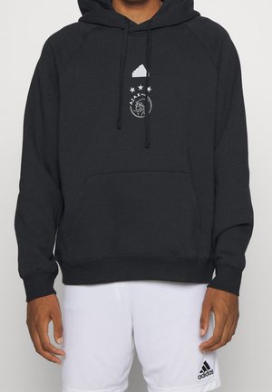 Mężczyzna w czarnej bluzie z kapturem Adidas z logo Ajax i białych krótkich spodenkach Adidas, stojący z ramionami swobodnie opuszczonymi wzdłuż ciała.