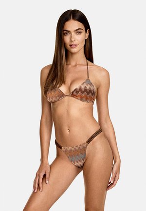 Donna che indossa un bikini marrone e arancione con motivo a zigzag e spalline laterali intrecciate, in piedi su uno sfondo chiaro e uniforme.