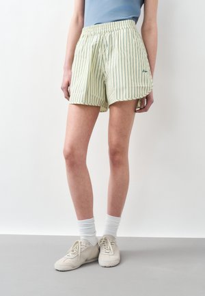 Persoană purtând pantaloni scurți cu dungi verticale în galben deschis și verde, un top albastru, șosete albe și adidași bej, stând în picioare lângă un perete simplu.