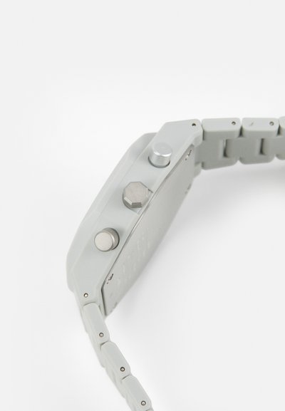 Γκρίζο πλαστικό ρολόι με ορθογώνια θήκη, τρία μεταλλικά κουμπιά και ευέλικτο λουράκι από αντίστοιχους γκρίζους συνδέσμους. Λείος υφής.