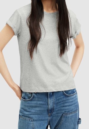 Femme portant un t-shirt gris clair uni à manches courtes et un jean bleu en denim, avec de longs cheveux bruns, debout les mains sur les hanches.