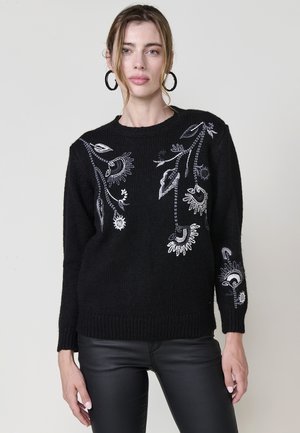 Suéter negro de punto con bordados florales en blanco y gris, escote redondo, y ribetes en el dobladillo y las mangas, combinado con pantalones negros.