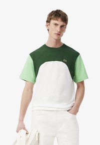 Camiseta con bloques de color que presenta una parte superior verde oscuro, mangas verdes claras y un cuerpo blanco. Hecha de algodón, con un cuello redondo y un logo.