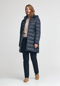 Marinblå pufferjacka med huva och horisontellt quiltad design, med dragkedja fram och en liten logotypdetalj. Kombineras med svarta byxor och bruna tofflor.