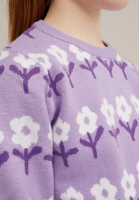 Pulover tricotat în lavandă, cu un model floral alb. Are un guler circular și un material texturat. Modelele florale sunt accentuate cu tulpini purpurii.