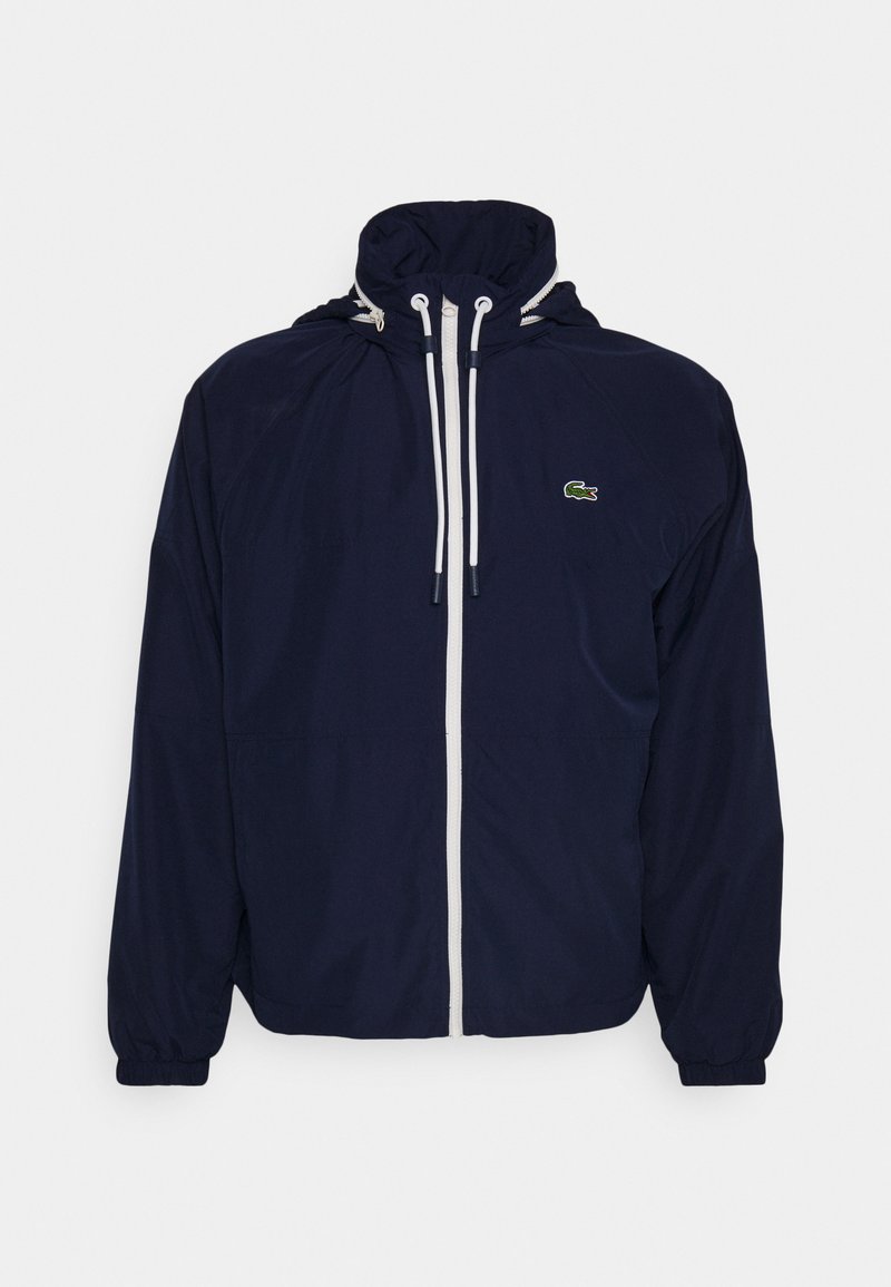 Giacca a vento blu navy con zip bianca e cordini, polsini elasticizzati, cappuccio e piccolo logo di coccodrillo verde sul lato destro del petto.