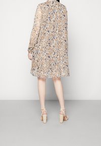 Blommönstrad klänning i beige och blå nyanser, med lång ärm, krage och A-linjeform, som matchas med ljusa klackar.