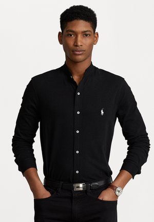 Zwarte lange mouwen shirt met een hoge kraag, witte knopen en een klein geborduurd logo op de linkerborst. Draagt met zwarte spijkerbroek en een riem.