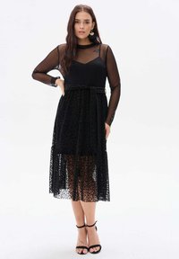Czarna sukienka z długimi przezroczystymi rękawami, dopasowanym gorsetem, kokardą w talii oraz przezroczystą spódnicą w wzór lamparta. Sandały na obcasie w czarnym kolorze.