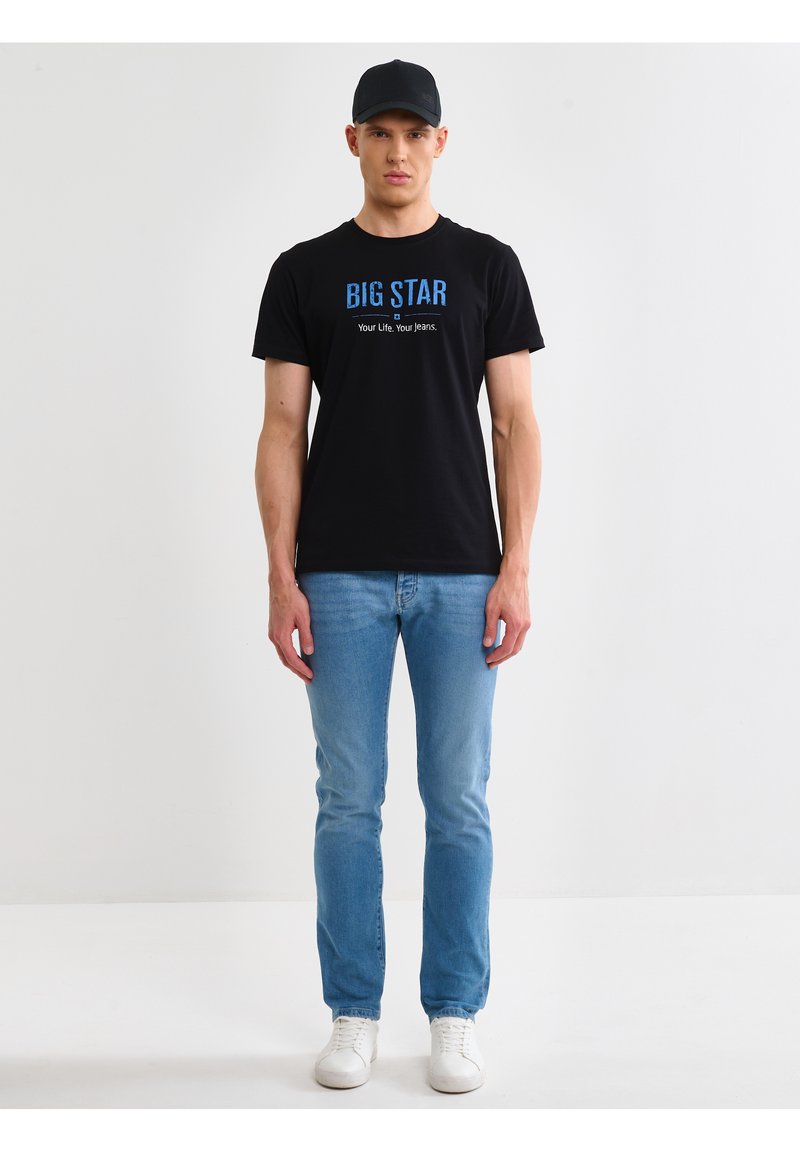 Muškarac u crnoj kapi, crnoj majici s natpisom "BIG STAR", svijetloplavim trapericama i bijelim tenisicama, stoji ispred jednostavne bijele pozadine.