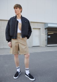 Veste bomber noire, shorts beiges, chaussures noires sans lacets et chaussettes blanches à rayures noires. La veste possède une fermeture éclair et des poches.
