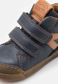 Chaussure en cuir bleu marine avec des accents marron, dotée de deux sangles Velcro réglables, surface texturée et semelle en caoutchouc avec motif de grip.