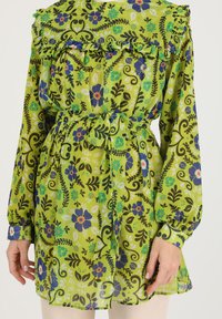 Green floral-patterned blouse with frilled neckline, long sleeves, and a tied waist belt. Made from lightweight, semi-sheer fabric.