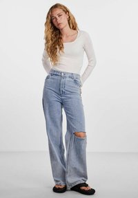 Top bianco a maniche lunghe a coste abbinato a jeans larghi azzurri con ginocchio strappato. Indossato con sandali neri flat.