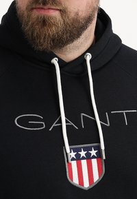 Zwarte hoodie met witte trekkoorden, voorzien van het "GANT"-logo en een schildpatch met rode en blauwe strepen en witte sterren. Gemaakt van katoen, sportief ontwerp.