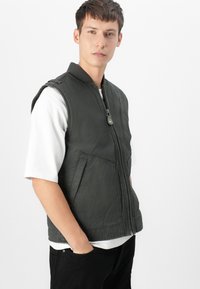 Chaleco sin mangas gris con cremallera en la parte delantera, dos bolsillos frontales y cuello acanalado, llevado sobre una camiseta blanca. Tejido suave con un acabado mate.