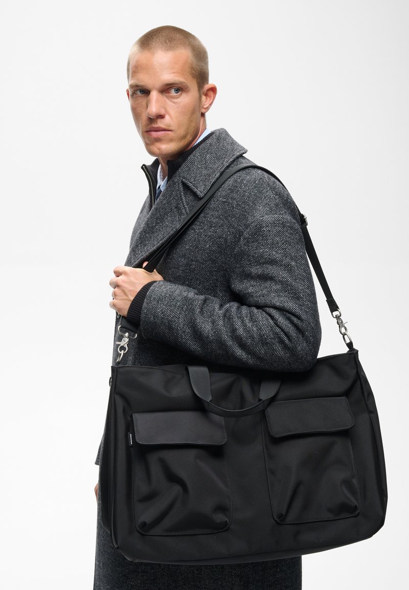 Homme portant un manteau gris à motif chevrons, portant un grand sac cabas noir avec des poches avant sur l'épaule, sur fond blanc.