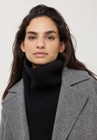 Cappotto grigio in misto lana con spalle strutturate, indossato sopra un dolcevita nero a coste. Texture liscia con dettagli di cucitura minimi.