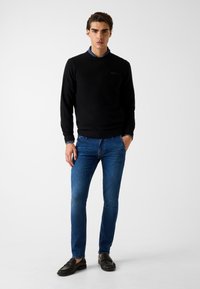 Czarny sweter z okrągłym dekoltem, niebieskie jeansy i czarne mokasyny. Prosty design z fakturowanej tkaniny. Model stoi na jednolitym tle.