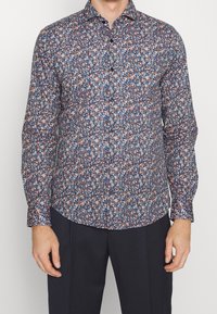 Man in een overhemd met lange mouwen en een dicht bloempatroon in blauw, wit en oranje, gecombineerd met donkere broek.