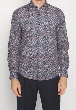 Uomo che indossa una camicia a maniche lunghe con una densa fantasia floreale nei colori blu, bianco e arancione, abbinata a pantaloni scuri.