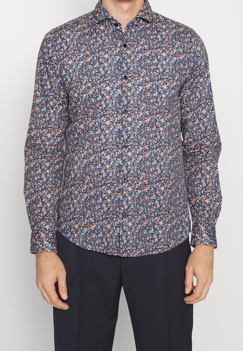 Man in een overhemd met lange mouwen en een dicht bloempatroon in blauw, wit en oranje, gecombineerd met donkere broek.
