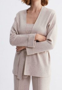 Beige stickad cardigan med V-ringning, långa ärmar och bälte i midjan, matchad med en topp och byxor i samma färg. Mjuk textur synlig.
