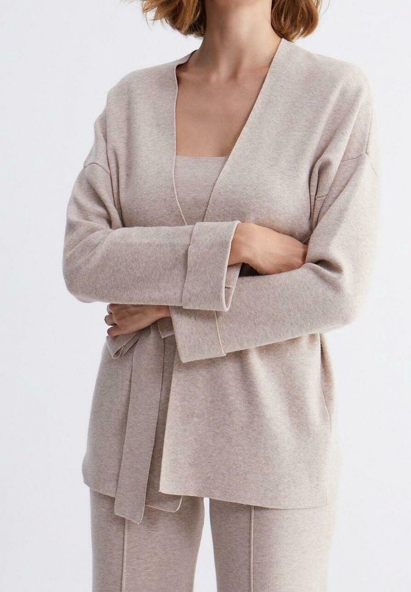 Beige stickad cardigan med V-ringning, långa ärmar och bälte i midjan, matchad med en topp och byxor i samma färg. Mjuk textur synlig.