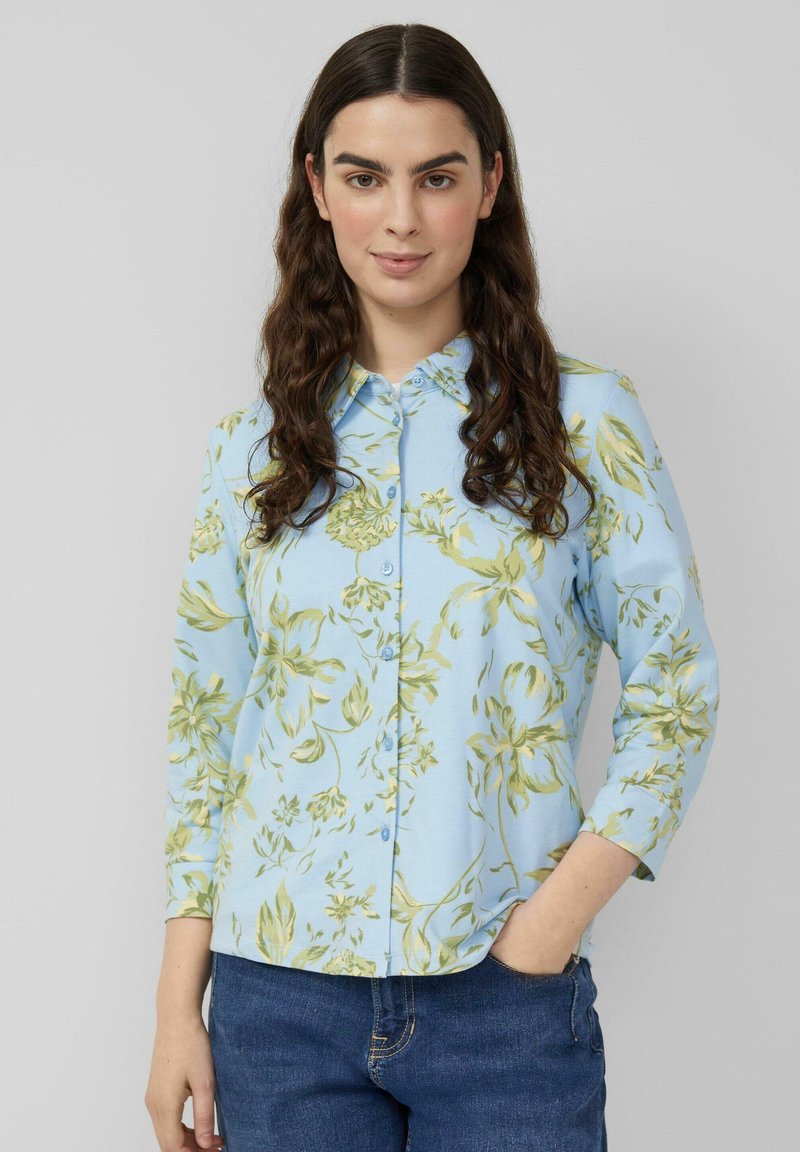 Mujer con cabello largo y oscuro, vestida con camisa azul claro con botones y patrón de flores amarillas, y jeans azules, con la mano en el bolsillo, fondo neutro.