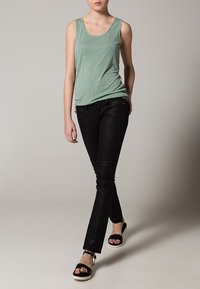 Top sin mangas verde menta, jeans skinny negros, sandalias negras con correas en el tobillo, modelo de pie con un fondo neutro.