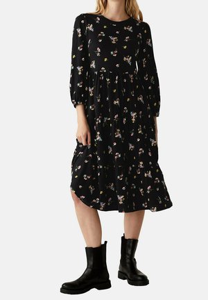 Femme portant une robe midi noire à motifs floraux avec des manches trois-quarts et des bottines noires épaisses, debout devant un fond uni.