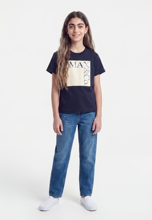 Fille aux longs cheveux portant un t-shirt noir Max&Co., un jean bleu et des chaussures blanches sans lacets, debout devant un fond blanc.