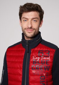 Gilet imbottito rosso con cuciture a trama, maniche blu navy e colletto alto. Presenta un logo e le coordinate stampate in blu sulla parte anteriore.