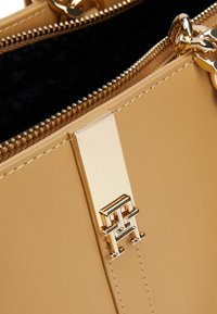 Sac à main couleur sable avec un extérieur en cuir lisse, des ferrures dorées, un logo accentué, et une ouverture zippée avec une doublure intérieure noire.