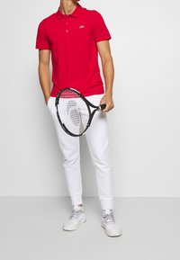 Red polo shirt with a collar and logo, paired with white sweatpants. Holding a tennis racket. Footwear is light gray athletic shoes.