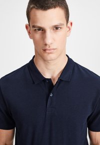 Polo shirt bleu marine en tissu texturé, avec une patte de boutonnage à trois boutons et un logo discret sur la poitrine gauche.